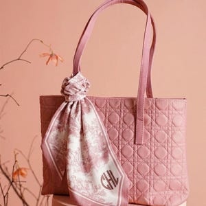 May include: A pink tote bag with a quilted pattern and matching handles. A decorative scarf with a landscape design and the initials "CHL" is tied to the handle. The bag is set against a soft pink background.