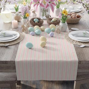 May include: A festive Easter table setting with a pink and green striped table runner. The table is decorated with pastel-colored eggs, floral arrangements, and decorative nests. White plates and napkins are set for dining.