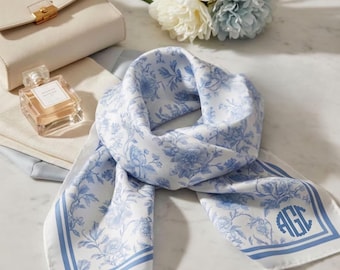Peut inclure: Un foulard carré en soie à fond blanc avec un motif floral bleu, une bordure bleue et le monogramme AGC imprimé dans un cadre circulaire. Le foulard est plié et disposé avec un sac à main crème et un flacon de parfum en verre à proximité.
