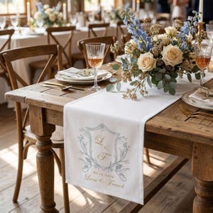 May include: A wooden table set for a wedding reception, featuring a white table runner with a custom crest design. The runner is adorned with floral arrangements, glassware, and place settings. The crest includes the initials "L" and "F", the date 05.16.2026, and the names "Lena & Franck".