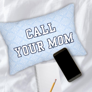May include: A light blue decorative pillow with the text "CALL YOUR MOM" in white and navy blue. A black smartphone, a spiral-bound notebook, and a pencil are also visible.