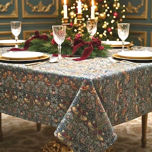 May include: A square tablecloth featuring a dense, intricate tapestry-style pattern of birds, flowers, and foliage in shades of blue, green, red, and gold. The fabric drapes over a table set with white plates, clear glasses, and a centerpiece.