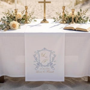 May include: White linen table runner with a light blue and gold crest design, floral accents, and the names "Lena & Franck" with the date 05.16.2026. The table is set for a wedding ceremony with candles and flowers.
