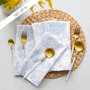 Può includere: Un set di quattro tovaglioli bianchi con un motivo floreale blu, disposti con cucchiai e forchette dorate. Le posate hanno manici bianchi. I tovaglioli sono su un sottobicchiere intrecciato. Il testo "SET OF 4" è visibile.