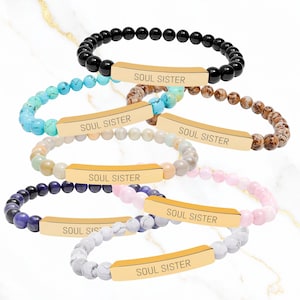May include: A collection of beaded bracelets in various colors, including black, turquoise, brown, cream, purple, pink, and white. Each bracelet features a gold-colored rectangular bar with the words "SOUL SISTER" engraved on it. The bracelets are displayed on a white marble surface.