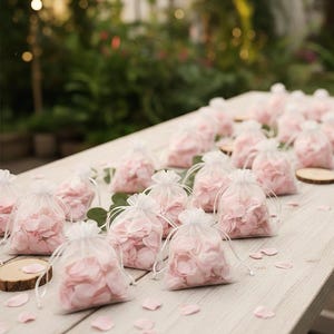 May include: Several small, transparent drawstring bags filled with pale pink flower petals, displayed on a light-coloured wooden surface. Scattered petals and small wooden discs enhance the decorative arrangement, possibly for a wedding.
