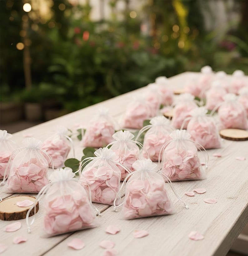 May include: Several small, transparent drawstring bags filled with pale pink flower petals, displayed on a light-coloured wooden surface. Scattered petals and small wooden discs enhance the decorative arrangement, possibly for a wedding.