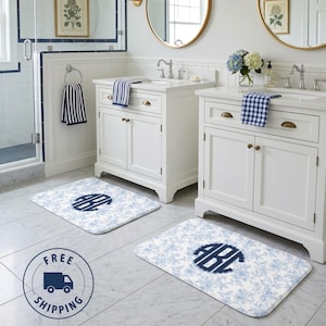 May include: Two rectangular bathroom mats with a blue floral pattern and a navy blue monogram. The mats are in front of white bathroom vanities with matching towels and decor. The bathroom has white walls and a marble floor.