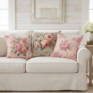May include: Three floral cushion covers with rose patterns in pink and green hues are arranged on a white sofa. The cushions have a vintage, romantic style, adding a touch of elegance to the living room.