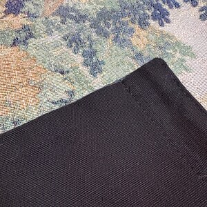 May include: Close-up of a black, textured fabric with a woven pattern. The fabric is a dark, solid color and appears to be a corner of a larger item. The background features a tapestry with floral and foliage patterns in shades of blue, green, and beige.