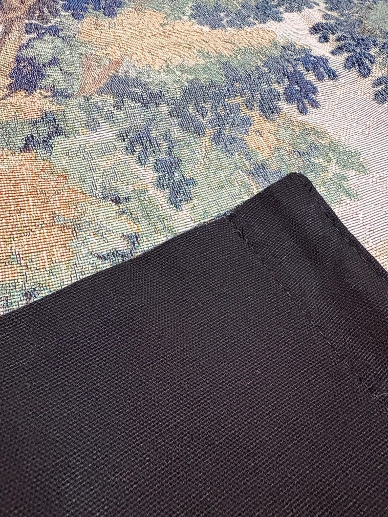 May include: Close-up of a black, textured fabric with a woven pattern. The fabric is a dark, solid color and appears to be a corner of a larger item. The background features a tapestry with floral and foliage patterns in shades of blue, green, and beige.