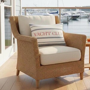 May include: A wicker armchair with a cream-colored cushion and backrest. A rectangular pillow with the words "YACHT CLUB" in red lettering is on the chair. The chair is on a wooden deck overlooking a marina.