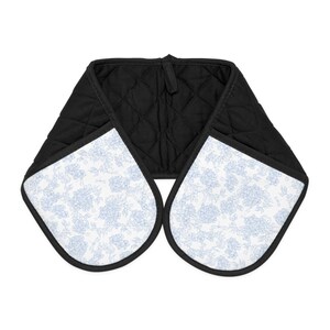 Puede incluir: Una manopla doble para horno con tela acolchada negra en el interior y exterior blanco con un patr&oacute;n floral azul claro. La manopla tiene dos bolsillos redondeados conectados por una tira central, con bordes rematados en tela negra.