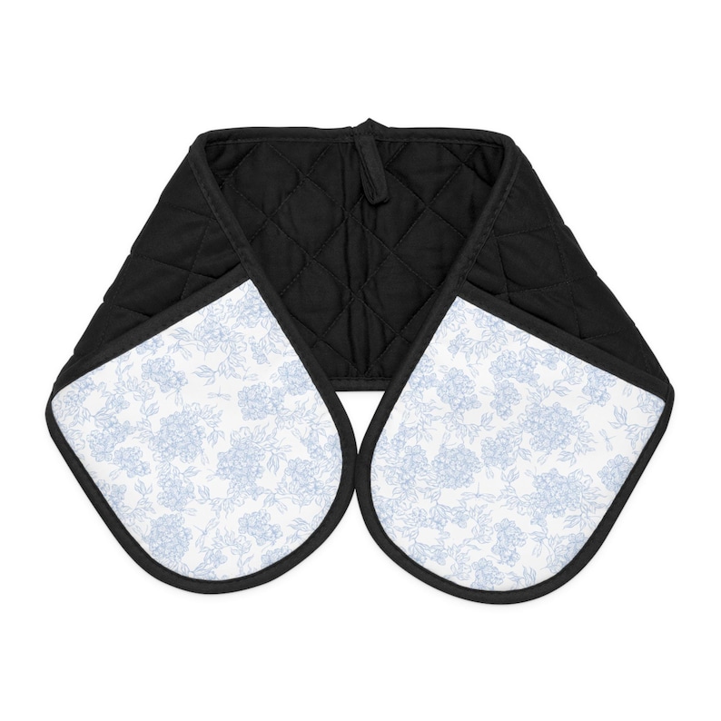 Puede incluir: Una manopla doble para horno con tela acolchada negra en el interior y exterior blanco con un patr&oacute;n floral azul claro. La manopla tiene dos bolsillos redondeados conectados por una tira central, con bordes rematados en tela negra.