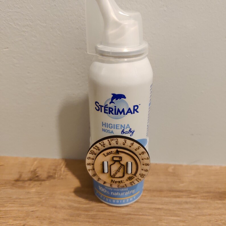 May include: A white bottle of St&eacute;rimar nasal hygiene spray for babies, with a blue dolphin logo. A wooden reminder with numbers and text is attached to the bottle. The text on the bottle reads "HIGIENA NOSA baby".