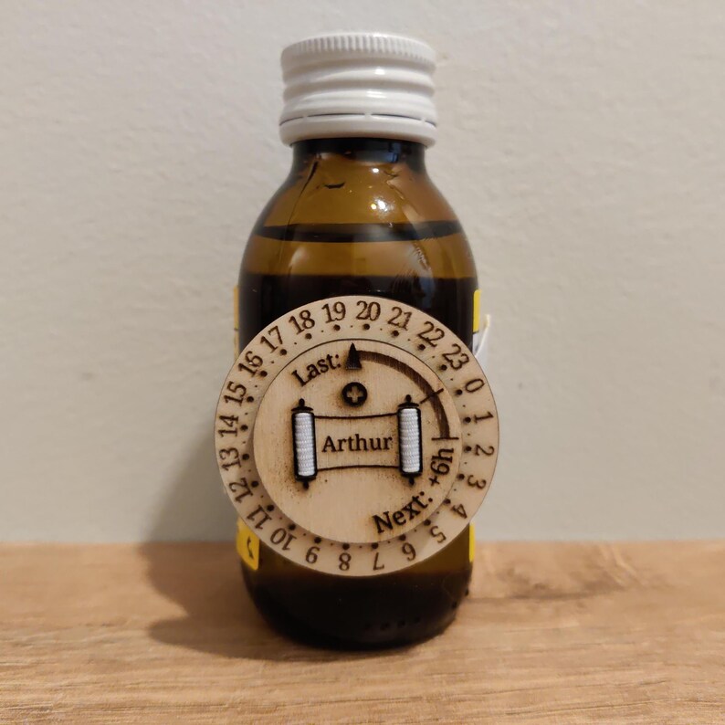 May include: A brown glass bottle with a white cap and a wooden tag. The tag has a circular design with numbers, the name "Arthur", and the words "Last" and "Next".