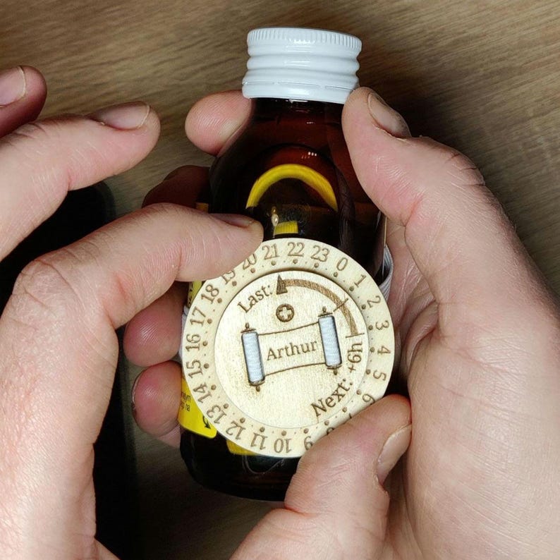 May include: A brown bottle with a white cap is held by two hands. A wooden dial with numbers and the name "Arthur" is attached to the bottle. The dial has the words "Last" and "Next" on it.