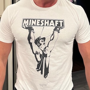 May include: A white short-sleeved t-shirt featuring a black graphic print of a person in a tank top and cap holding up a rectangular sign that reads MINESHAFT. Below the main graphic, smaller text reads THE MINESHAFT 835 WASHINGTON STREET NEW YORK, N.Y. 10014.