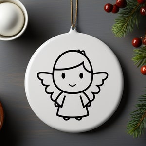 Op de afbeelding: Witte, ronde kerstversiering met een zwarte omtrekillustratie van een cartoon engel. De engel heeft vleugels, een halo en een lachend gezicht. Een gouden draad is bevestigd om op te hangen. Achtergrond met dennentakken.
