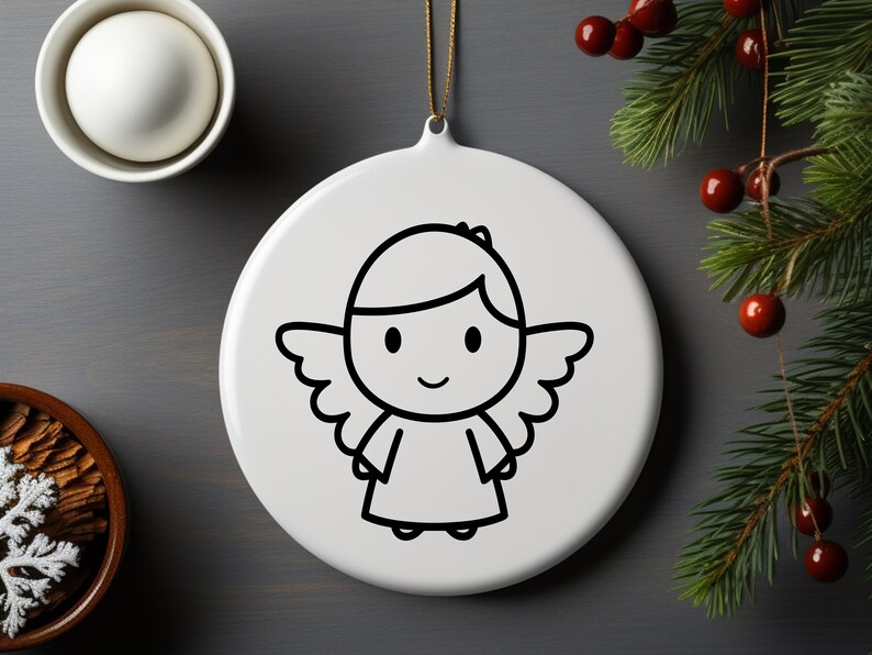 Op de afbeelding: Witte, ronde kerstversiering met een zwarte omtrekillustratie van een cartoon engel. De engel heeft vleugels, een halo en een lachend gezicht. Een gouden draad is bevestigd om op te hangen. Achtergrond met dennentakken.