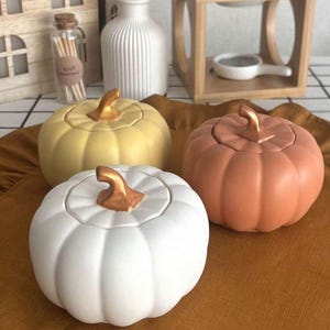 Puede incluir: Tres recipientes de cerámica con forma de calabaza en blanco, amarillo y naranja, cada uno con un tallo dorado. Las calabazas están dispuestas sobre una superficie marrón, lo que sugiere un tema de otoño o cosecha.
