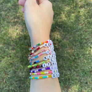 May include: A person's wrist wearing a stack of colorful beaded bracelets with letter beads spelling out the words "GET, IF, KING, STAY, IT, WAS, OUR, YOU, DES"