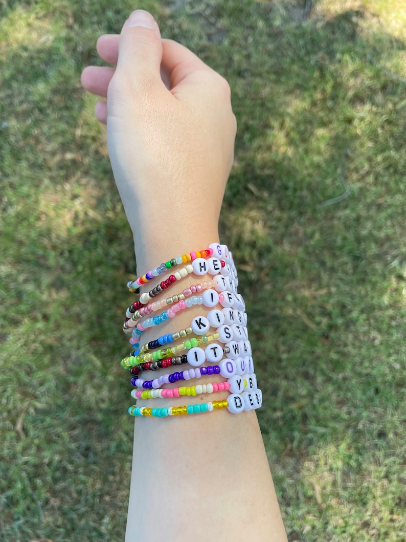 May include: A person's wrist wearing a stack of colorful beaded bracelets with letter beads spelling out the words "GET, IF, KING, STAY, IT, WAS, OUR, YOU, DES"