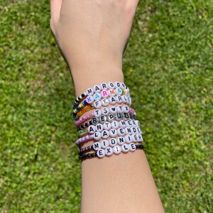May include: A stack of colorful beaded bracelets with white beads that spell out words like "MAROON", "KARMA", "FEARLESS", "TS", "REPUTATION", "ANTI-HERO", "LAVENDER", "MIDNIGHT", and "EXILE".