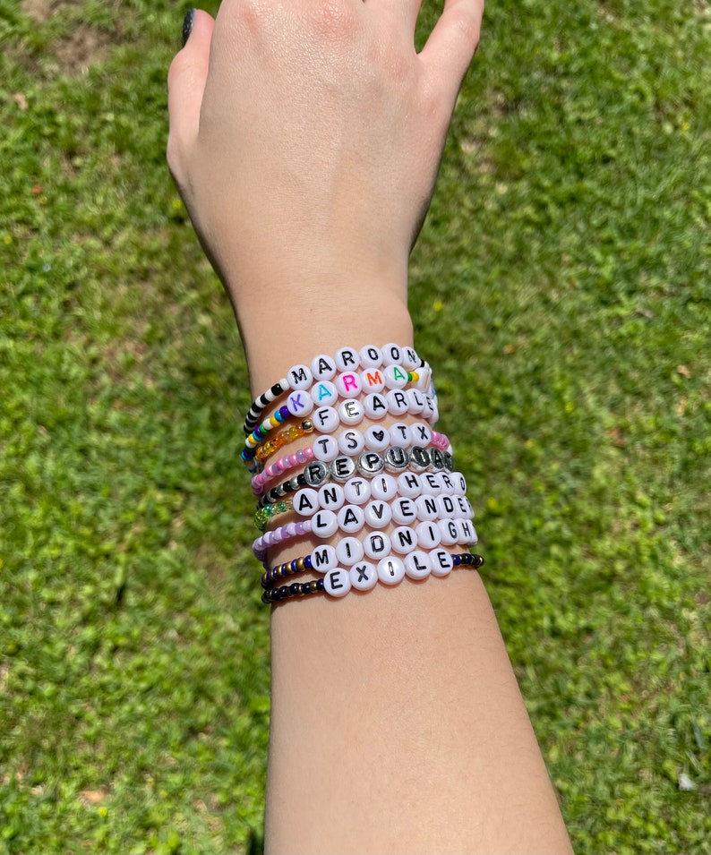 May include: A stack of colorful beaded bracelets with white beads that spell out words like "MAROON", "KARMA", "FEARLESS", "TS", "REPUTATION", "ANTI-HERO", "LAVENDER", "MIDNIGHT", and "EXILE".