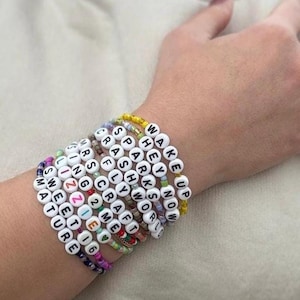 May include: A collection of beaded bracelets spelling out words and phrases like "Wake Up," "Hey Now," and "Sweet 16." The bracelets feature white letter beads on colorful string, creating a personalized and trendy accessory.