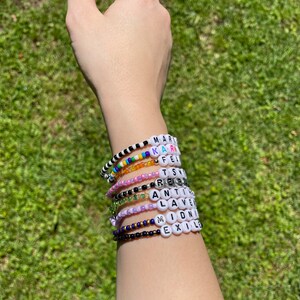 May include: A person's wrist with multiple beaded bracelets. The bracelets are made with white letter beads that spell out the words "MAR", "KAR", "FE", "TS", "RE", "ANT", "LAV", "IDN", "EXIL", and "M".