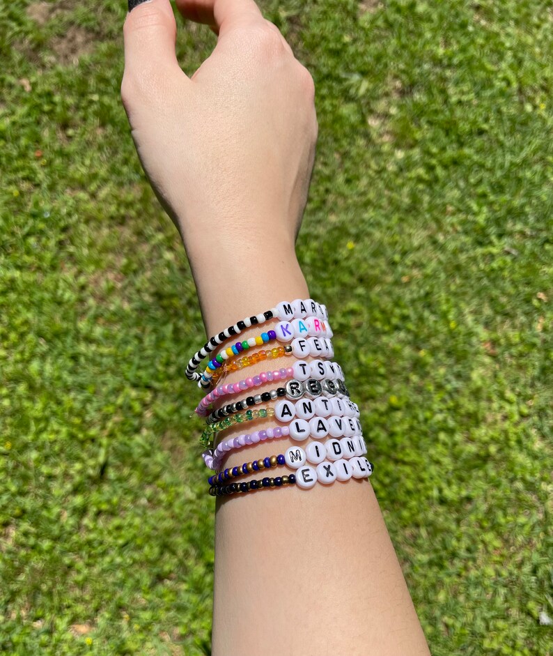 May include: A person's wrist with multiple beaded bracelets. The bracelets are made with white letter beads that spell out the words "MAR", "KAR", "FE", "TS", "RE", "ANT", "LAV", "IDN", "EXIL", and "M".