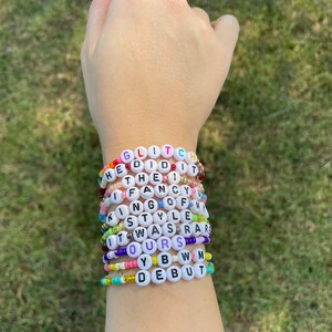 May include: A stack of colorful beaded bracelets with white letter beads spelling out the phrases "GLITCH", "HE DID IT", "THE I", "IF FANCY", "KING OF", "STYLE", "IT WAS RARE", "OURS", "YBW", and "DEBUT".