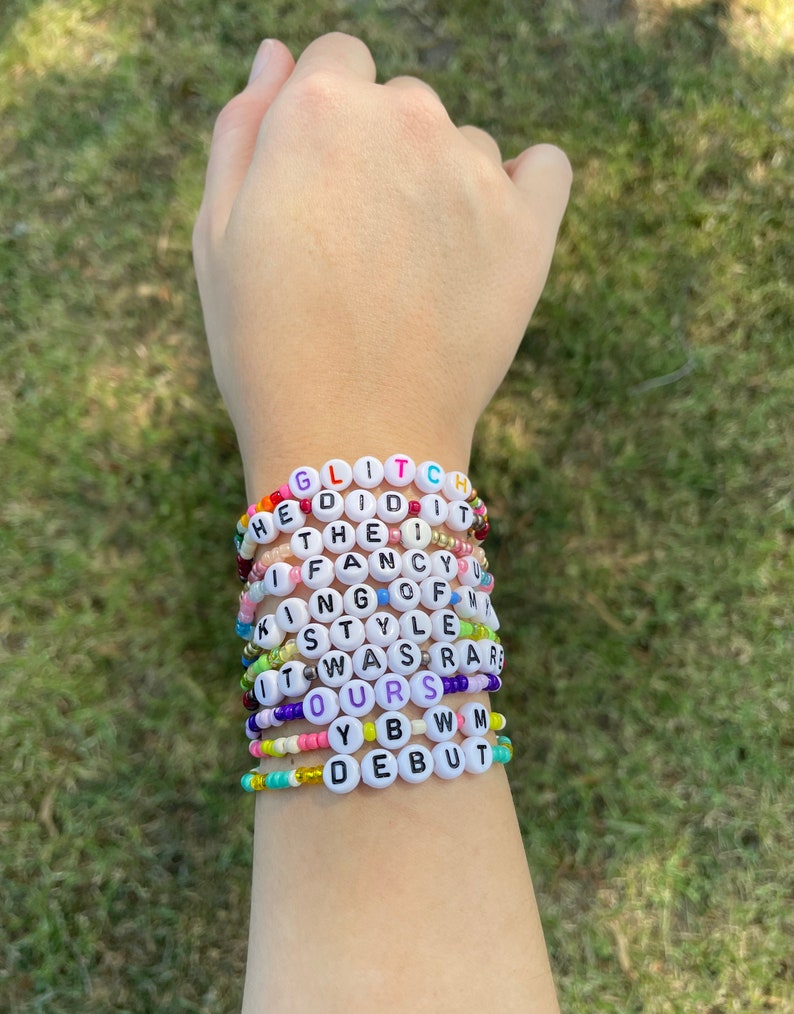 May include: A stack of colorful beaded bracelets with white letter beads spelling out the phrases "GLITCH", "HE DID IT", "THE I", "IF FANCY", "KING OF", "STYLE", "IT WAS RARE", "OURS", "YBW", and "DEBUT".