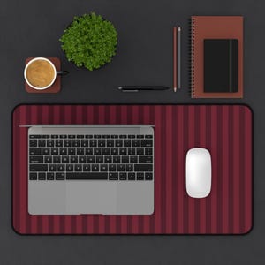 May include: A silver laptop and a white computer mouse sit on a rectangular desk mat featuring a dark red and black vertical striped pattern. Nearby are a brown spiral notebook, a black journal, two pencils, a black pen, and a coffee mug.