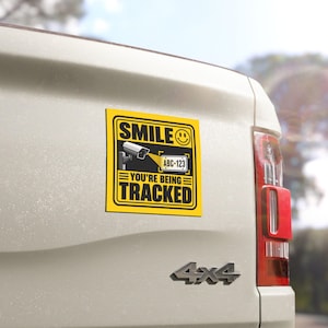 Puede incluir: Una pegatina cuadrada amarilla con borde negro que muestra un gráfico de cámara de seguridad, un icono de cara sonriente y una matrícula con el texto ABC-123. El texto SMILE YOU'RE BEING TRACKED aparece en letras negras mayúsculas.