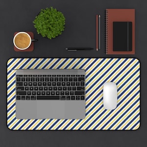 May include: A rectangular desk mat featuring a diagonal striped pattern in navy blue, yellow, and white. A silver laptop with a black keyboard is centered on the mat, accompanied by a white wireless computer mouse positioned to the right side.