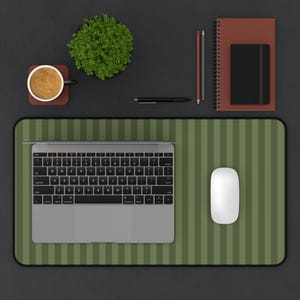 May include: A rectangular desk mat featuring a vertical olive green and dark green striped pattern. The mat is positioned under a silver laptop and a white computer mouse. The surface is flat with rounded corners and a black border trim.
