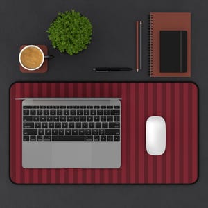 May include: A rectangular desk mat featuring a dark red and black vertical striped pattern. The mat is positioned under a silver laptop with a black keyboard and a white computer mouse. The setup includes a coffee mug, a small potted plant, two pencils, a black pen, a spiral-bound notebook, and a small black journal.
