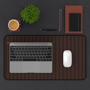 May include: A brown striped desk mat featuring a silver laptop and a white computer mouse. The workspace includes a cup of coffee on a coaster, a small green plant, two pencils, a black pen, a spiral-bound notebook, and a black journal.