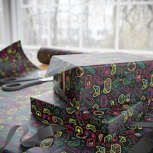 Peut inclure: Papier cadeau avec un motif répétitif de manettes de jeu vidéo et de consoles portables aux couleurs néon sur fond noir. Le papier recouvre deux boîtes rectangulaires, avec un rouleau et des ciseaux visibles à proximité.
