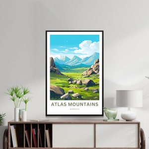 Op de afbeelding: Een ingelijste kunstdruk met een landschap van het Atlasgebergte in Marokko. Het kunstwerk toont een vallei met groen gras, grote rotsen en verre blauwe bergen onder een helderblauwe lucht met wolken. De tekst "ATLAS MOUNTAINS MOROCCO" staat onderaan.