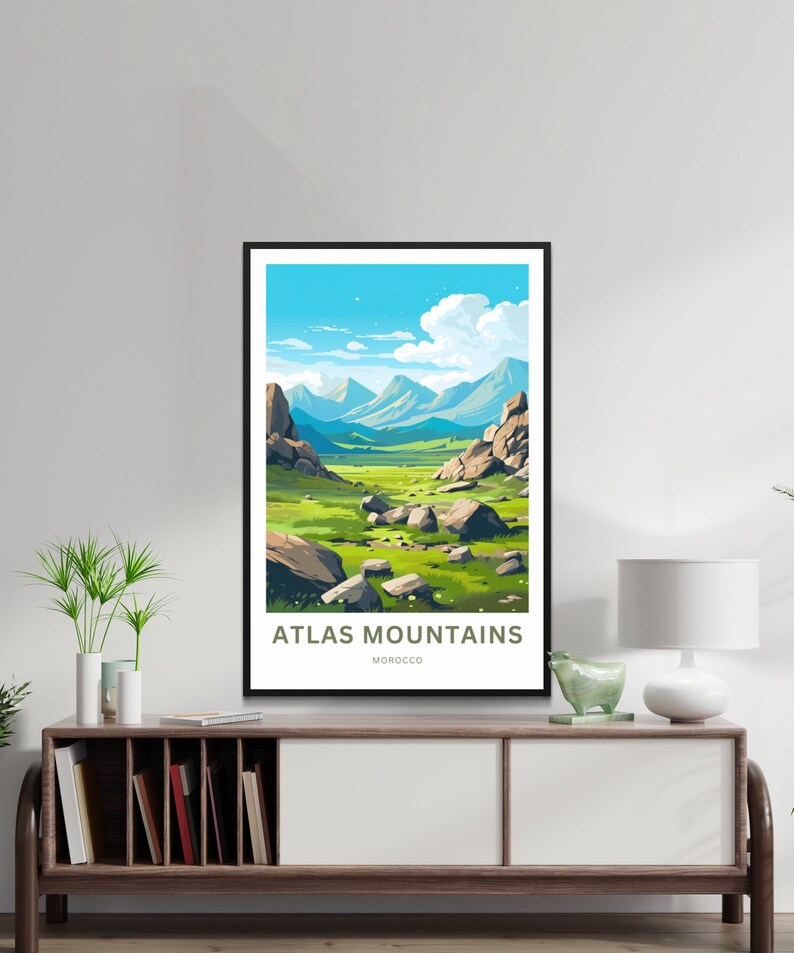 Op de afbeelding: Een ingelijste kunstdruk met een landschap van het Atlasgebergte in Marokko. Het kunstwerk toont een vallei met groen gras, grote rotsen en verre blauwe bergen onder een helderblauwe lucht met wolken. De tekst "ATLAS MOUNTAINS MOROCCO" staat onderaan.