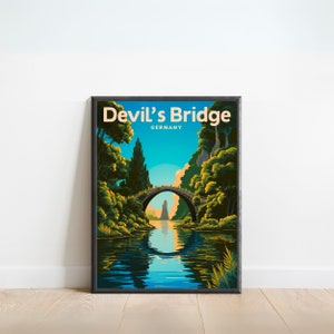 May include: A vintage travel poster with a black frame featuring a scenic view of Devil's Bridge in Germany. The poster has a blue sky, green trees, a stone bridge over a river, and the text "Devil's Bridge GERMANY" in a yellow and orange font.