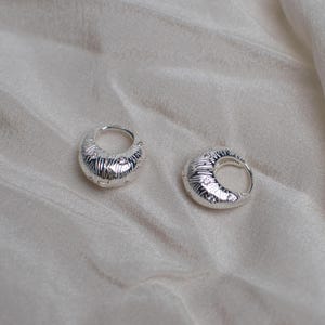 Peut inclure: Une paire de petites créoles argentées au design texturé en forme de croissant. La surface de chaque boucle d'oreille présente une série de fines lignes gravées rayonnant depuis le centre, créant une finition métallique détaillée sur le métal.