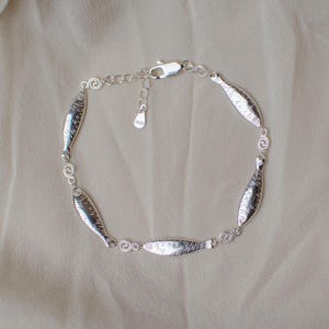 May include: A silver-toned bracelet featuring a series of small, stylized fish-shaped links connected by tiny circular spiral accents. The piece includes a lobster claw clasp and an adjustable chain extension with a small, dangling metal tag.