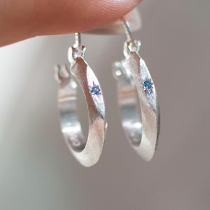 May include: A pair of silver hoop earrings with a brushed matte finish. Each hoop features a small, circular blue gemstone set into the center of the outer band. The earrings have a hinged closure mechanism at the top for secure attachment.