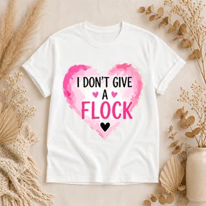 May include: White t-shirt with a pink heart graphic. The heart design features the text "I DON'T GIVE A FLOCK" in black and pink, with small heart accents. A casual, everyday wear tee.