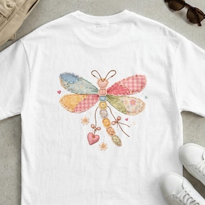 May include: White t-shirt featuring a colorful dragonfly design. The dragonfly is made of patchwork fabric in various pastel colors, including pink, blue, yellow, and green. The design includes small hearts and floral accents, creating a whimsical and artistic look.