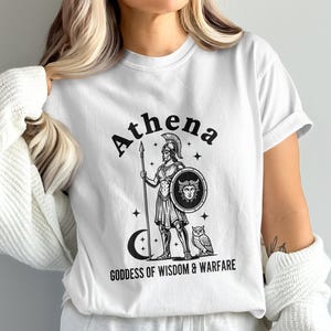 May include: White t-shirt featuring a black and white graphic of Athena, the Greek goddess of wisdom and warfare. The design includes Athena holding a spear and shield, with an owl and crescent moon. Text reads "Athena Goddess of Wisdom & Warfare."